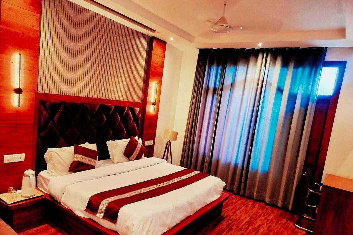 A modern hotel room with a large bed, wooden paneling, a red accent wall, and a view of the city through curtains.