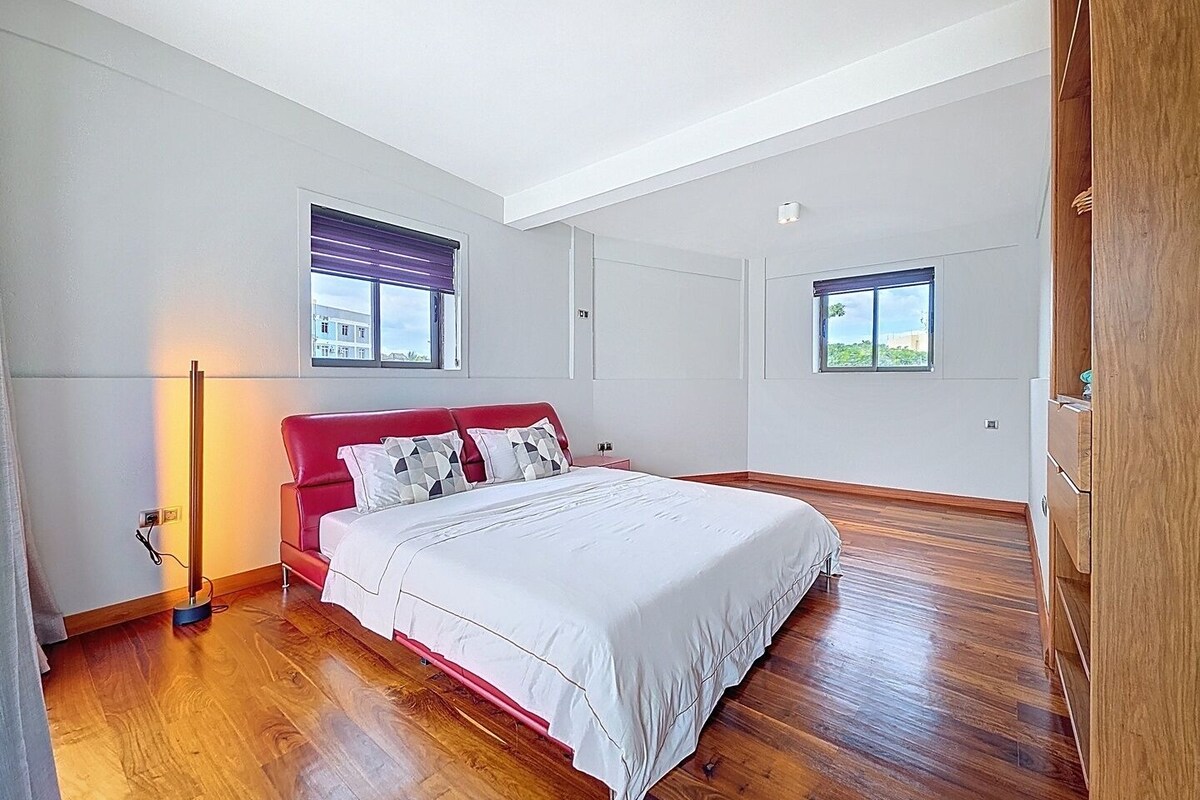 A bedroom with a red bed, white bedding, and two windows with blinds.