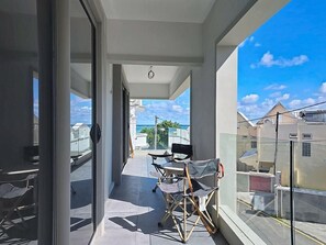 A balcony with a view of the sea, a table with chairs, and a glass door leading to the interior. - Blue Waves by dodo et Croissant (Blue Bay)