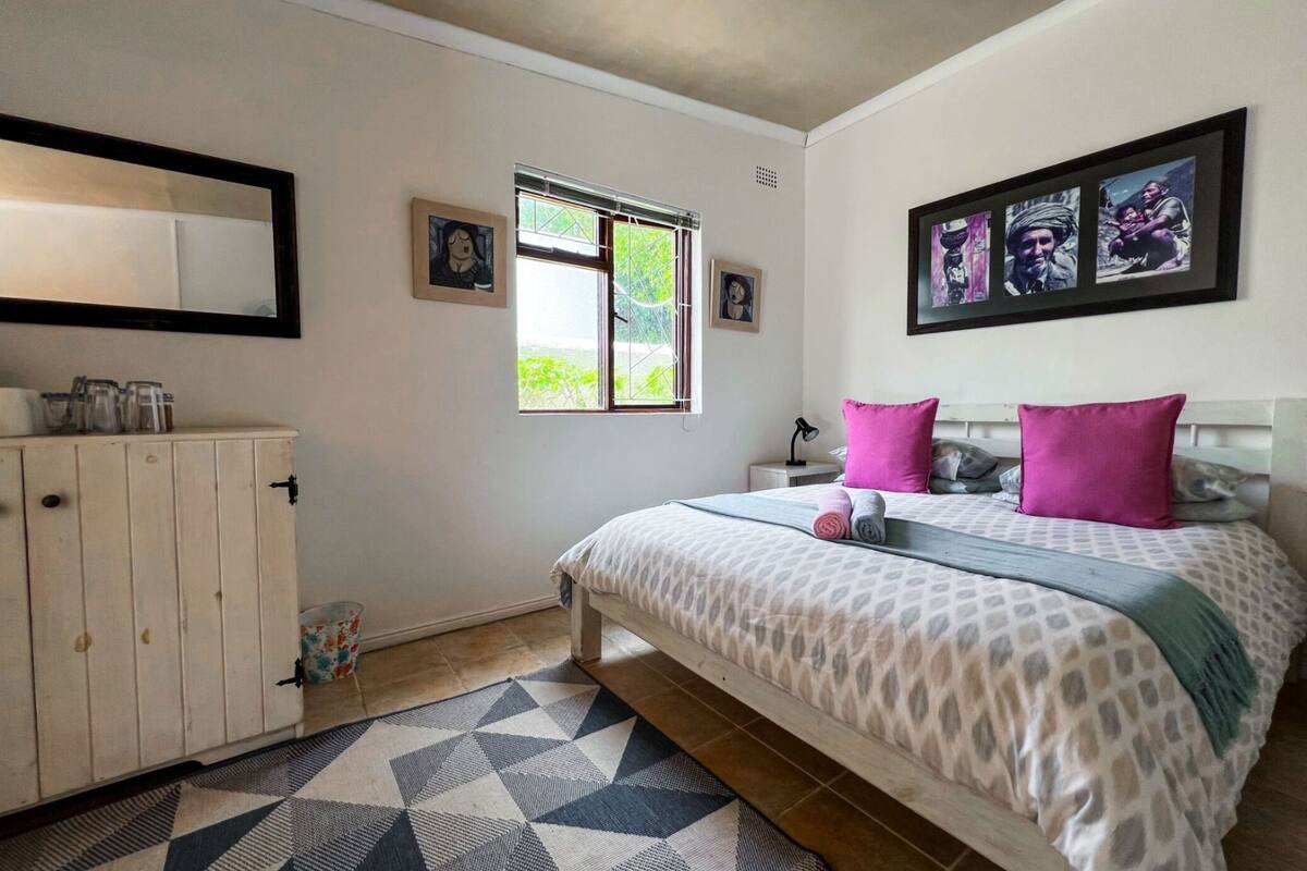 A bedroom with a bed, a nightstand, a dresser, a window with blinds, and wall art.