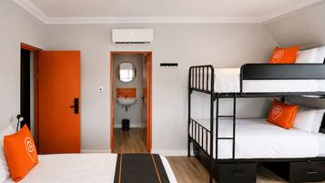 A hotel room with a bunk bed, a single bed, an orange door, a bathroom with a sink, and an air conditioning unit.