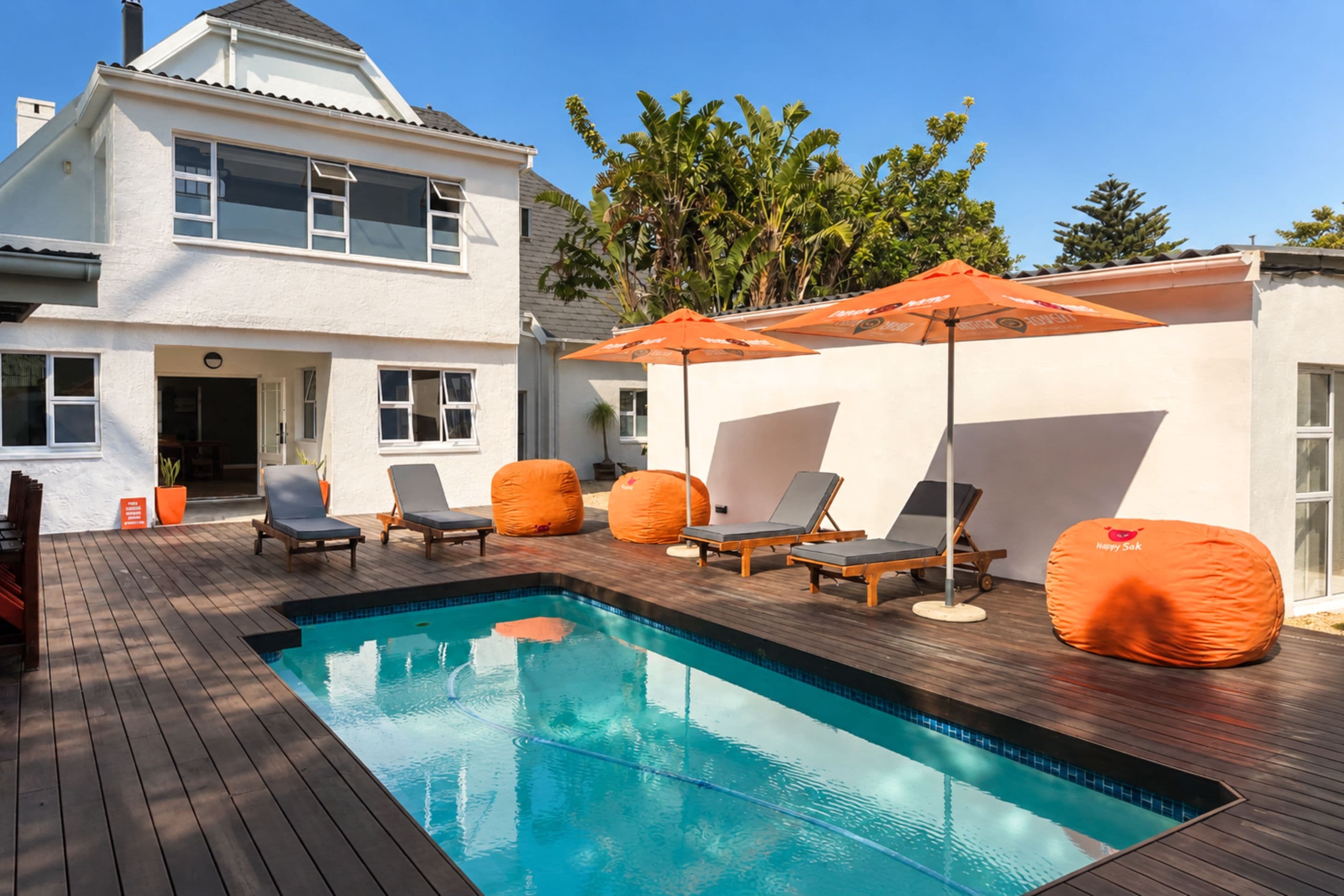 A swimming pool with lounge chairs and an umbrella, surrounded by a wooden deck and a white building.