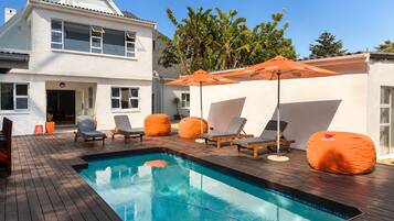 A swimming pool with lounge chairs and an umbrella, surrounded by a wooden deck and a white building.