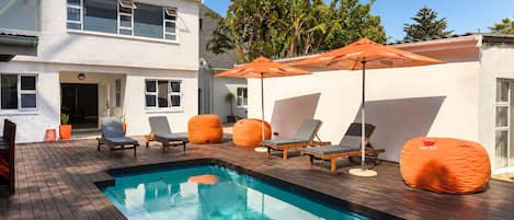 A swimming pool with lounge chairs and an umbrella, surrounded by a wooden deck and a white building.