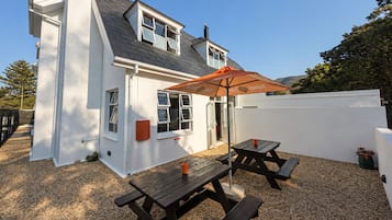 A white house with a grey roof, a wooden deck, and a picnic table with an umbrella.