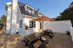 A white house with a grey roof, a wooden deck, and a picnic table with an umbrella.
