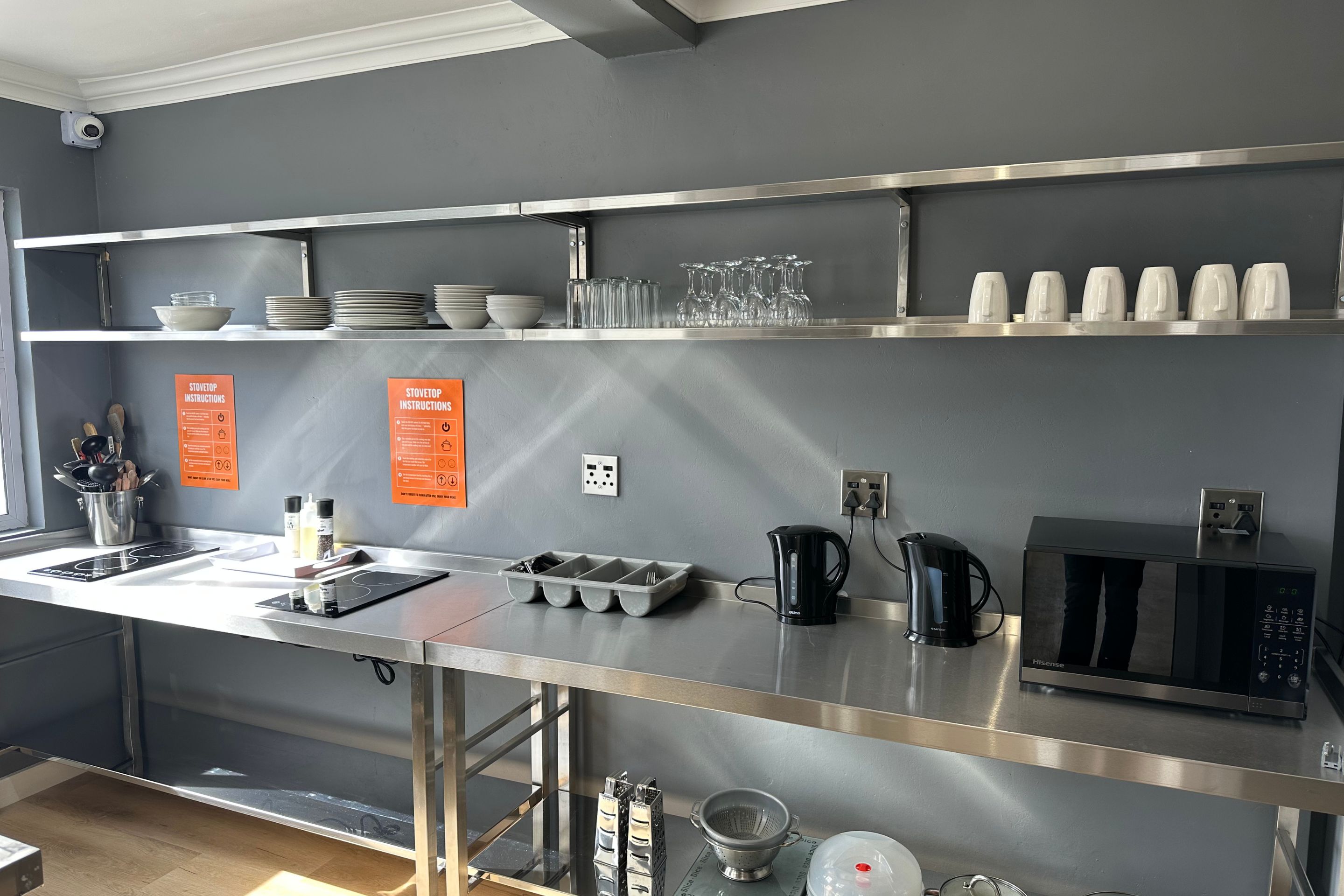 A modern kitchen with stainless steel appliances, including a microwave, kettle, and stove. There are shelves with dishes and glasses, and a sink with utensils.
