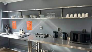 A modern kitchen with stainless steel appliances, including a microwave, kettle, and stove. There are shelves with dishes and glasses, and a sink with utensils.
