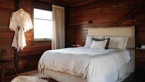 A wooden cabin bedroom with a bed, a hanging robe, a chair, and a small table with a vase.