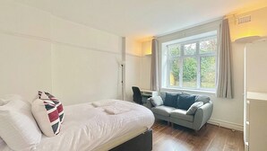 A bedroom with a bed, a grey sofa, a window with curtains, and a desk with a chair. - Finchley Ground Floor Apartment (London)