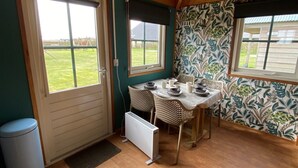 A compact kitchen with a dining table, two chairs, a radiator, and a window with a view of a grassy area. - Boerencamping Duinzoomhoeve - Hooiberghut Doré (Den Helder)
