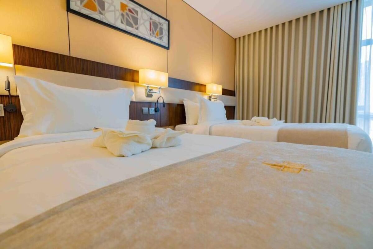Quarto de hotel com duas camas, cabeceira com iluminação embutida, uma janela grande com cortinas e uma obra de arte emoldurada na parede.