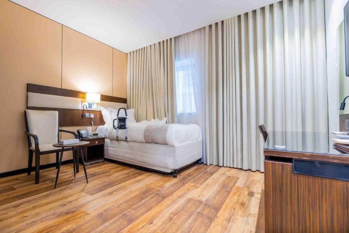 Quarto de hotel com cama, escrivaninha, cadeira, abajur e uma grande janela com cortinas.