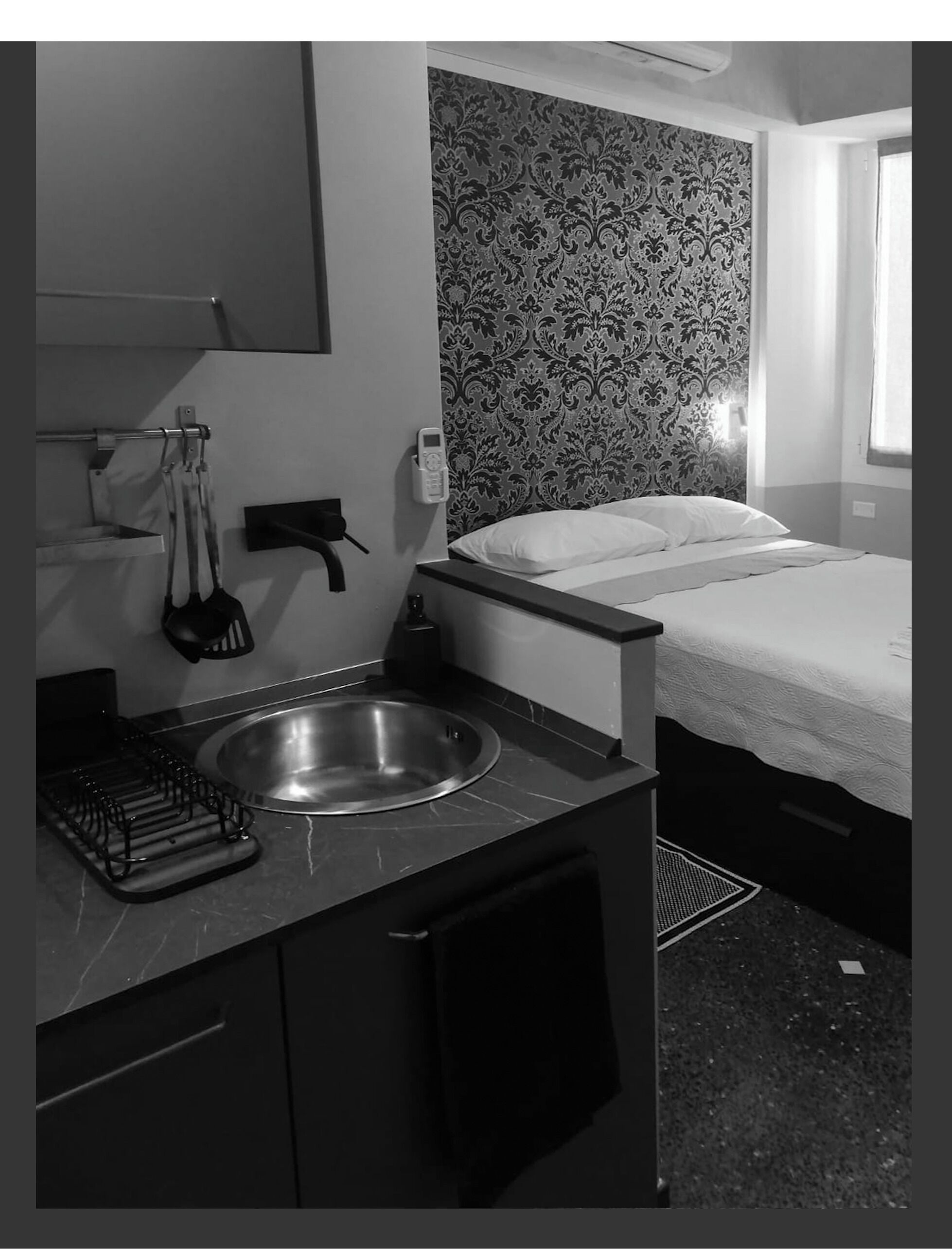 Habitación de hotel compacta con cama, una kitchenette pequeña con lavamanos y papel tapiz estampado.