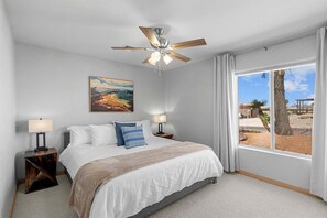 Un dormitorio con cama, ventilador de techo, un cuadro, ventana y vistas al exterior.