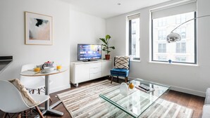 Een moderne woonkamer met een glazen salontafel, een televisie op een witte kast, een blauwe stoel en een groot raam met uitzicht op gebouwen.