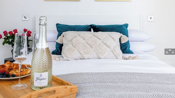 Hotelkamer met een bed, twee ingelijste abstracte schilderijen, een fles Freixenet Prosecco, een dienblad met croissants en bessen, en een vaas met rode bloemen.