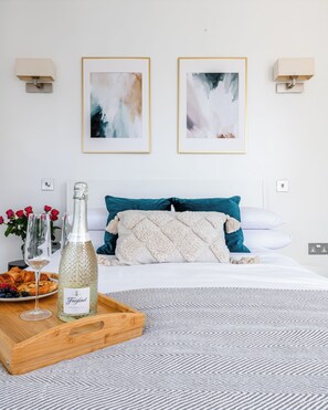 Habitación de hotel con una cama, dos cuadros abstractos enmarcados, una botella de Freixenet Prosecco, una bandeja con croissants y bayas, y un florero con flores rojas.