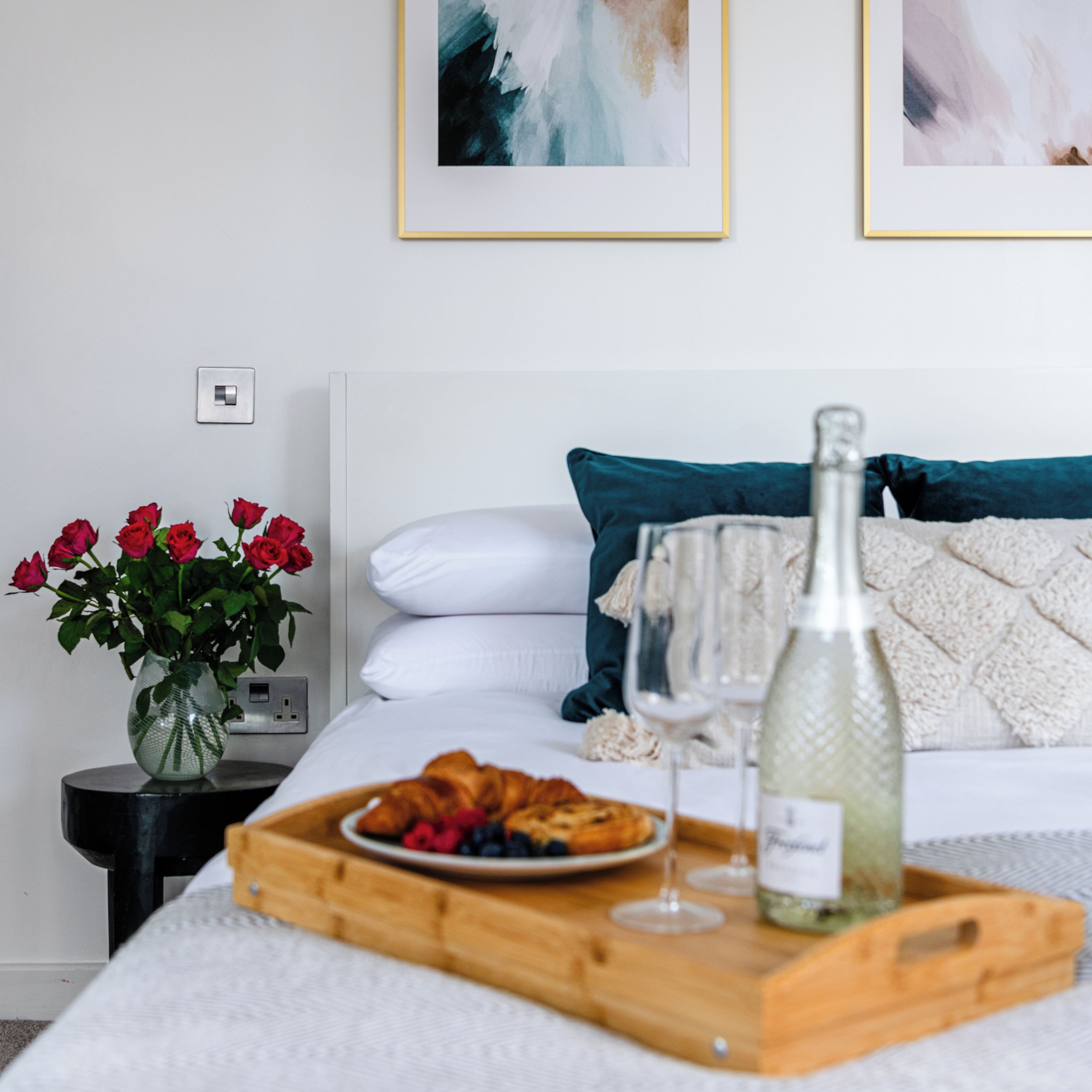 Une chambre à coucher bien rangée, avec un lit, une table de chevet ornée d’un vase de roses rouges, un plateau avec de la nourriture et une bouteille de vin, et deux tableaux abstraits accrochés au mur.