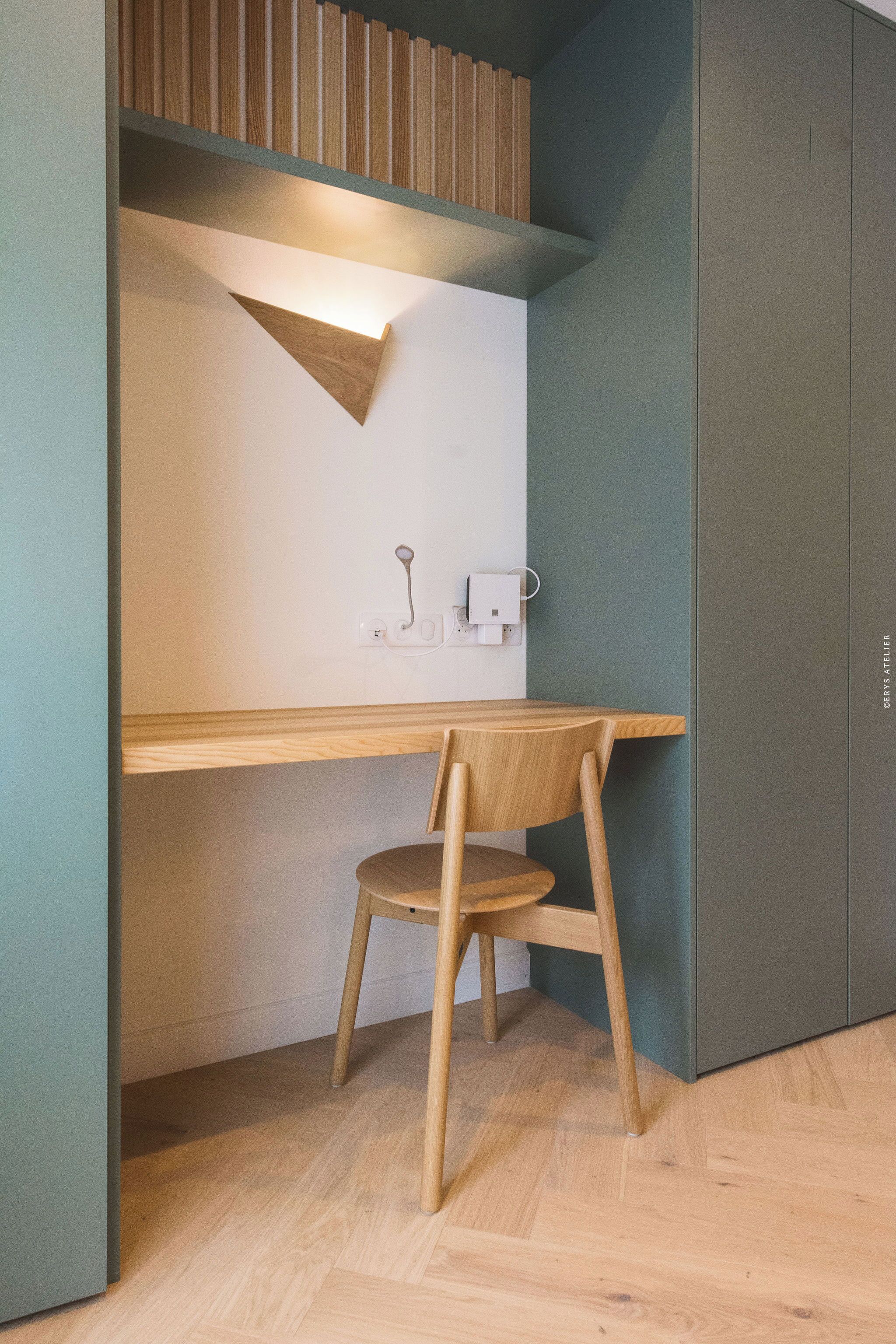 Un espacio de trabajo compacto con un escritorio y silla de madera, una lámpara fijada a la pared y un armario.