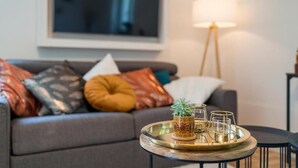 A modern living room with a grey sofa adorned with various cushions, a round coffee table with a tray holding a plant and glasses, and a floor lamp. - EG R City Wohnung Balkon nähe Salzburg (Freilassing)