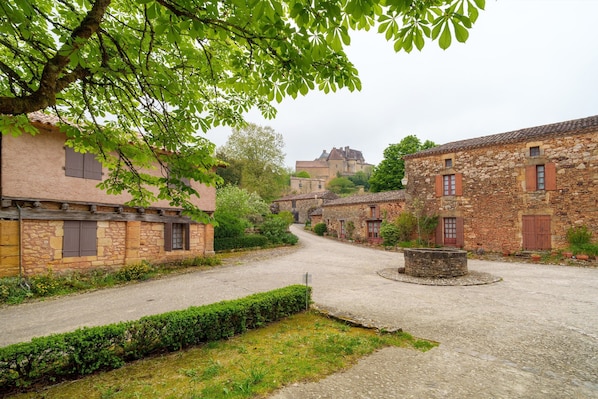 A cobblestone street in a historic village with stone buildings, a central roundabout, and a large castle in the background.