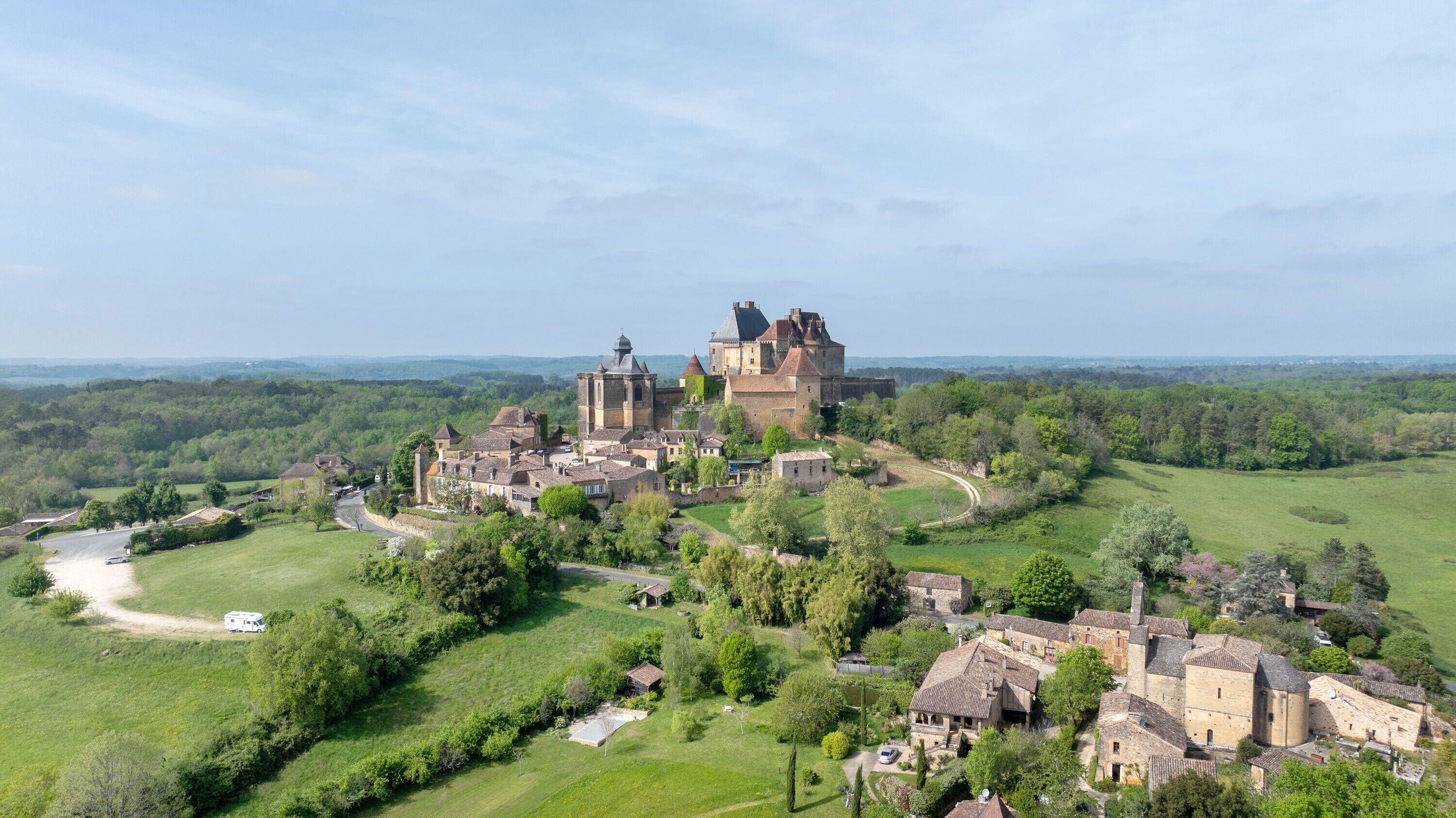 A historic castle complex with multiple buildings, surrounded by lush greenery and a clear sky.