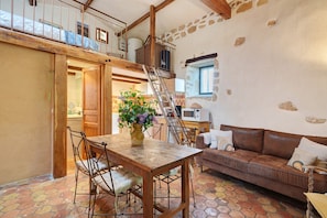 A rustic interior with a wooden dining table, a brown leather sofa, and a staircase leading to a loft.