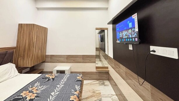 A modern bedroom with a bed, a wooden wardrobe, a TV mounted on the wall, and a small bedside table. - Devi Heritage (Mathura)