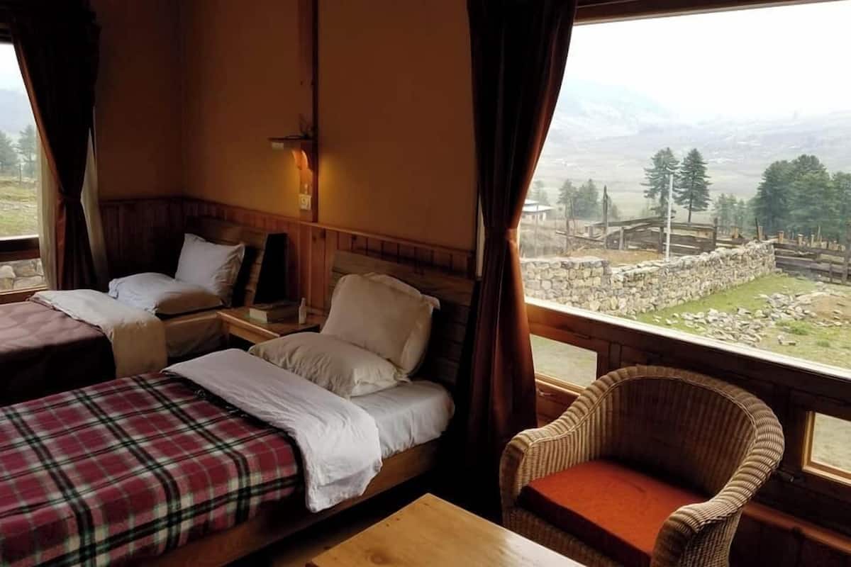 A cozy cabin room with two beds, a wicker chair, and a view of a mountainous landscape.