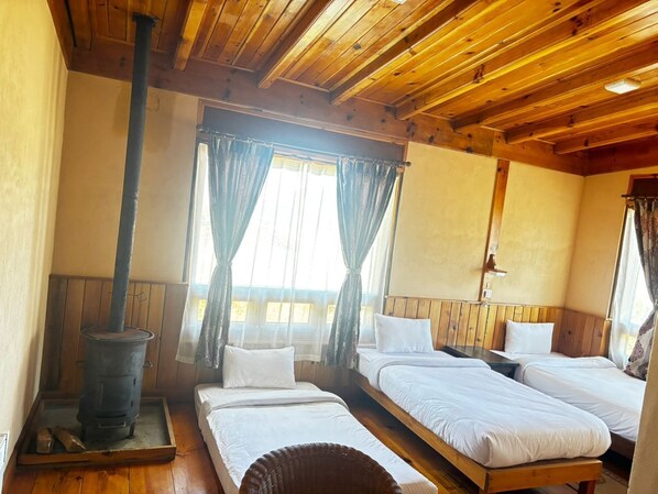A cozy room with wooden beams, a wood-burning stove, two beds, and large windows with curtains. - Kichu Resort Phobjikha (Wangdue Phodrang)
