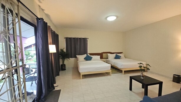 Quarto de hotel com duas camas de solteiro, uma mesinha, uma cadeira, um vaso de planta e vista pela janela com cortinas.