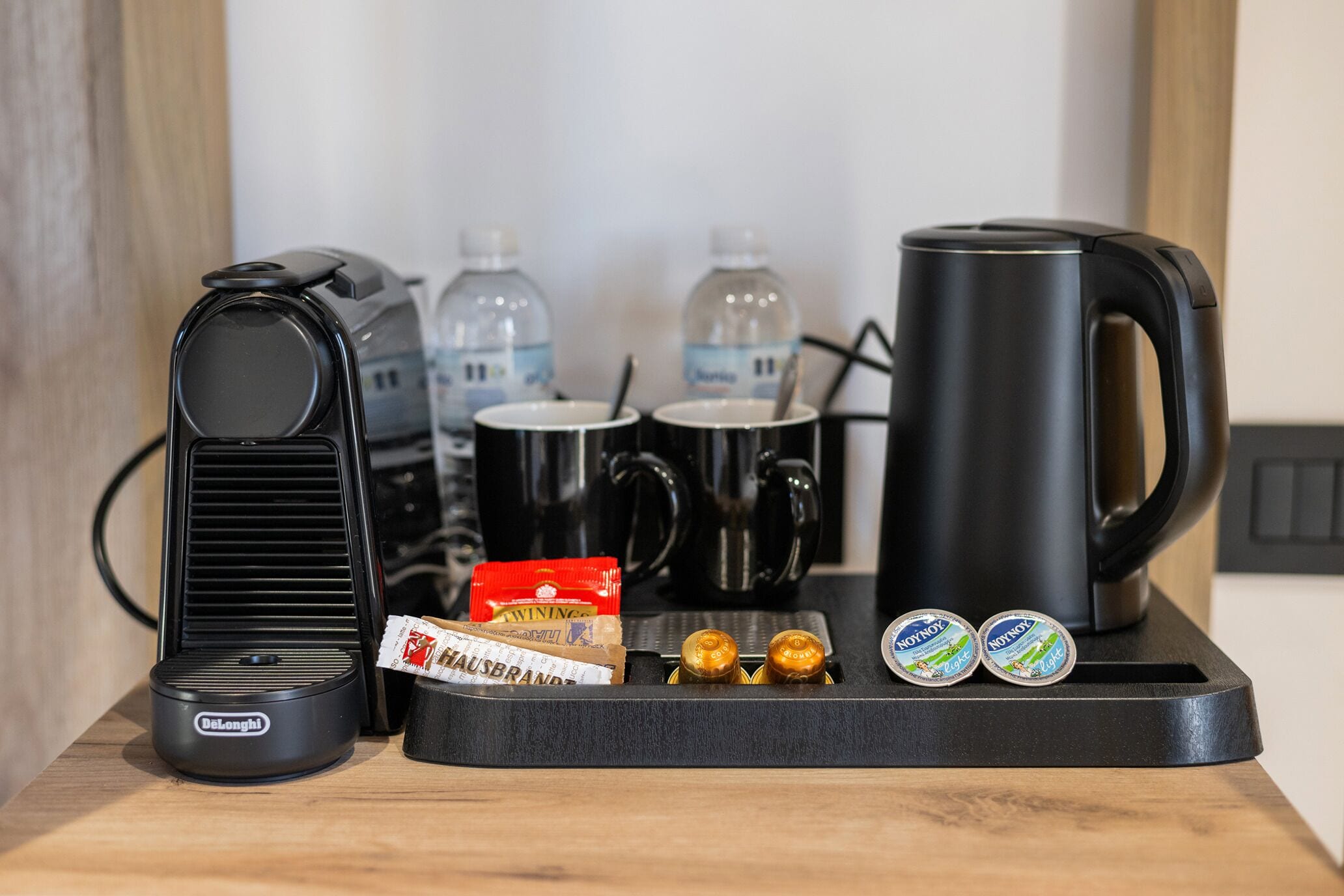 A coffee station with a Delonghi coffee maker, two black mugs, two water bottles, a red snack pack, and two containers of yogurt.