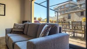 A grey sofa with patterned cushions in a room with large windows overlooking a courtyard with outdoor furniture and plants. - Tuscia Home Holidays - Rustic Elegance & Comfort (Civitella D'agliano)