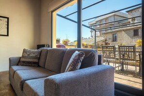 Um sofá cinzento com almofadas estampadas numa sala com grandes janelas com vista para um pátio com móveis de exterior e plantas.