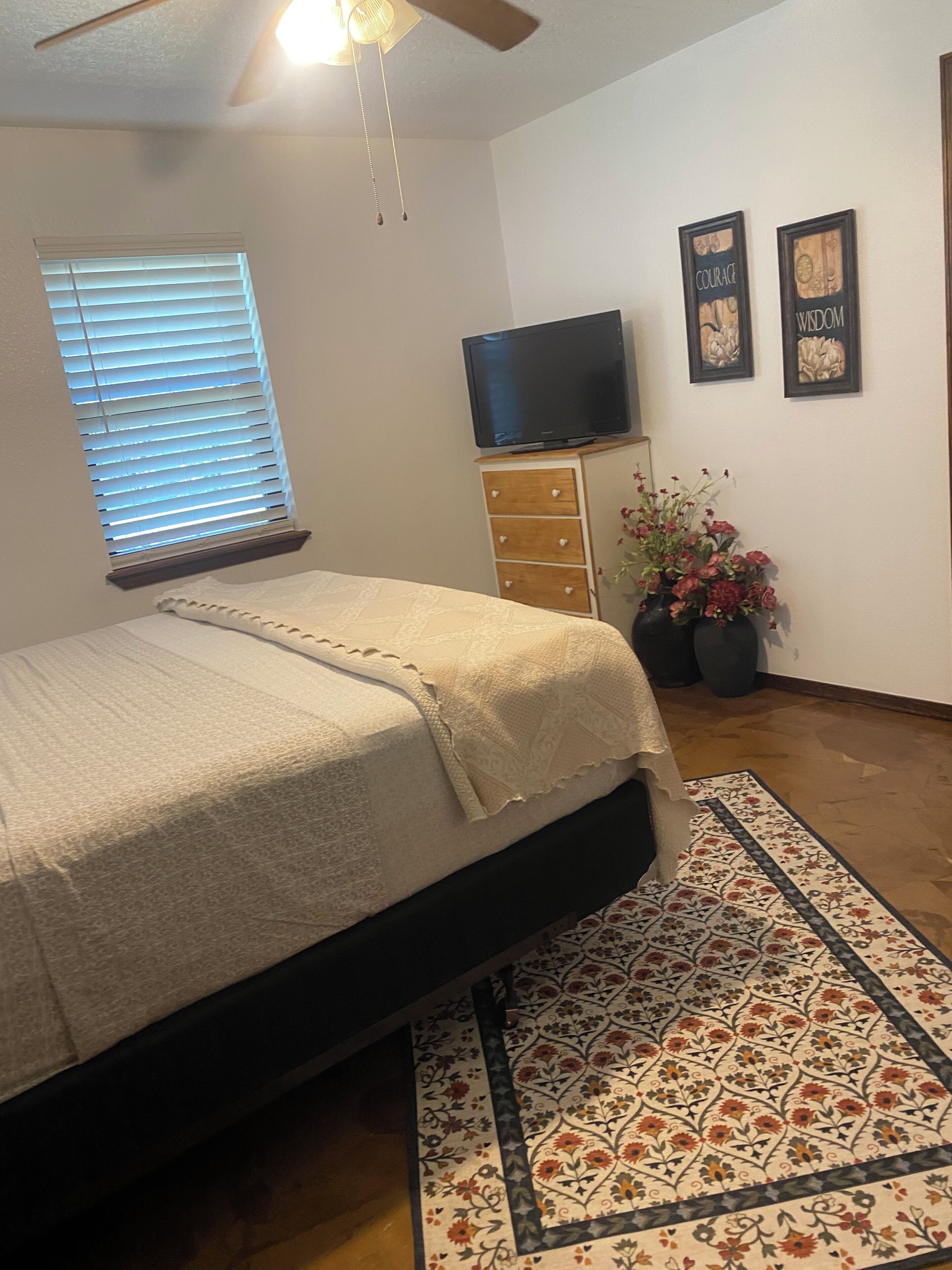 Un dormitorio con una cama, un televisor sobre una cómoda, una ventana con persianas y dos cuadros en la pared.