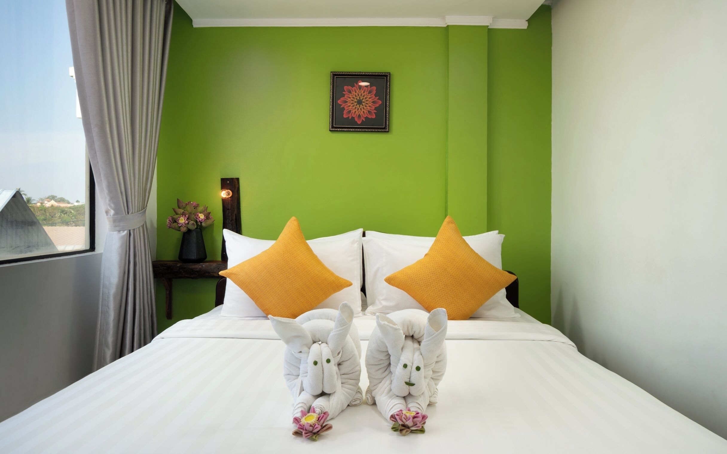 A hotel room with a green wall, a framed artwork, a bedside table with a vase of flowers, and two towels shaped like bunnies with flowers.