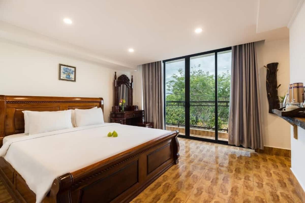 A hotel room with a large bed, a wooden headboard, a nightstand, a mirror, and a view of greenery through a large window.