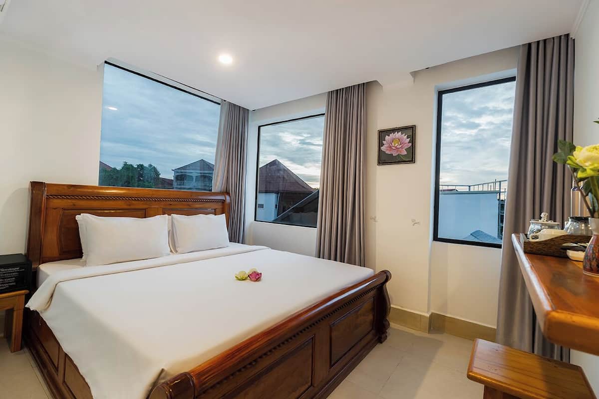 A hotel room with a large bed, two windows, a wooden headboard, and a bedside table with a vase of flowers.
