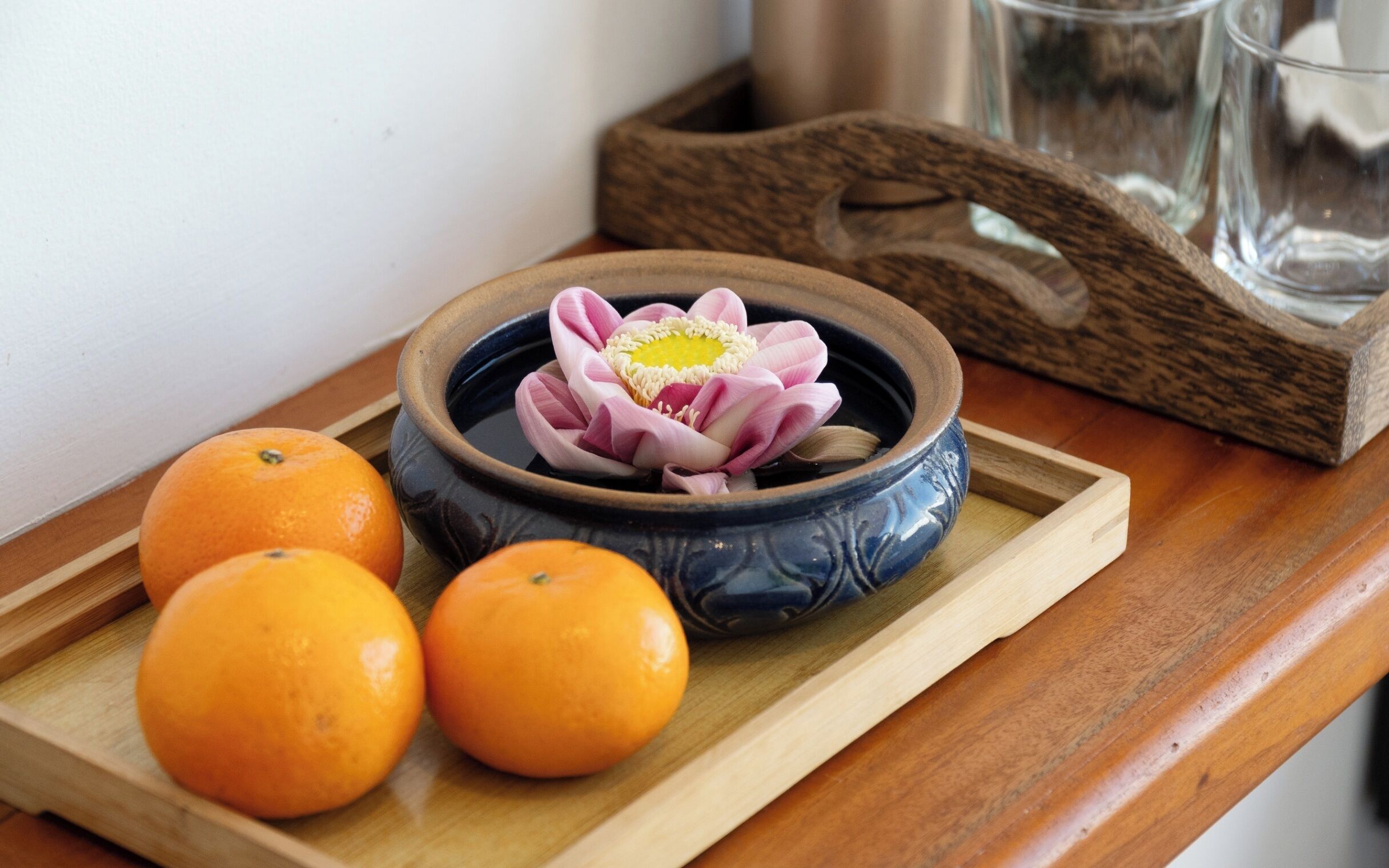 A wooden tray with a ceramic bowl containing a pink lotus flower, two oranges, and a glass.
