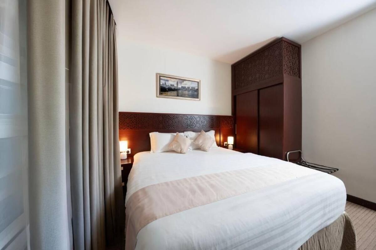 A hotel room with a large bed, two bedside tables with lamps, a dark wooden headboard, a framed picture on the wall, and a wardrobe.