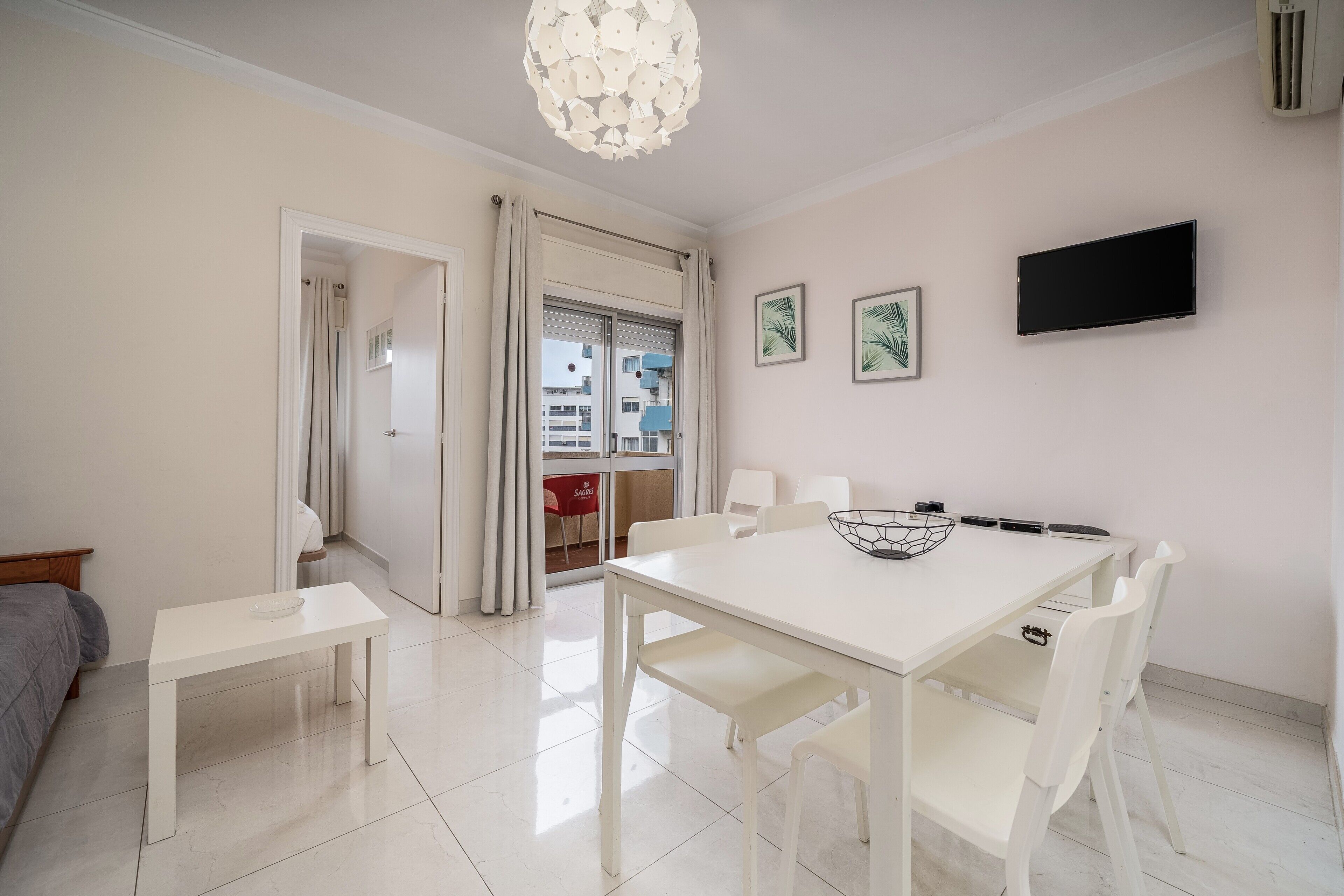 A dining area with a white table and chairs, a ceiling light fixture, and a flat-screen TV mounted on the wall.