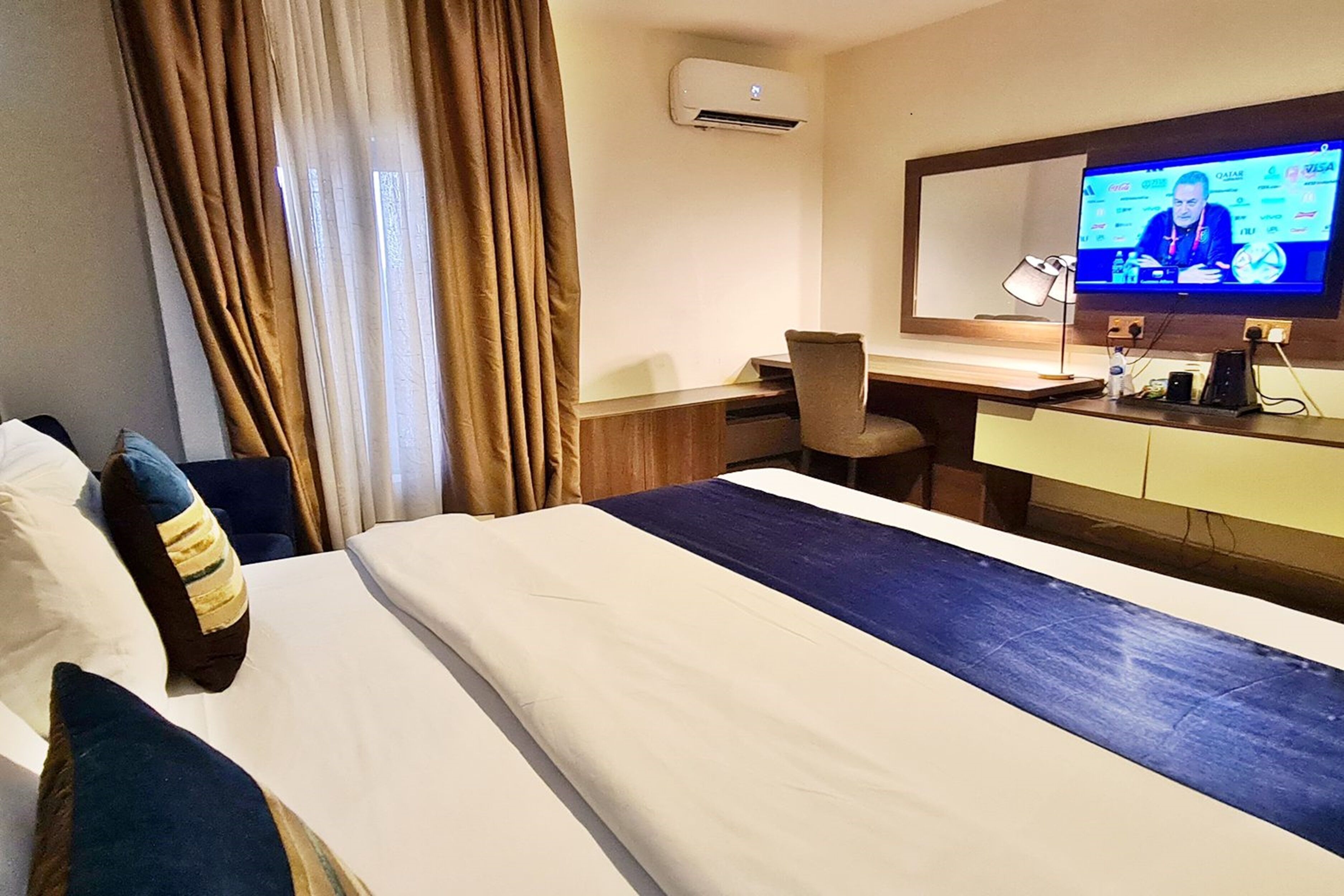 A hotel room with a bed, a desk, a chair, a TV, and an air conditioning unit.