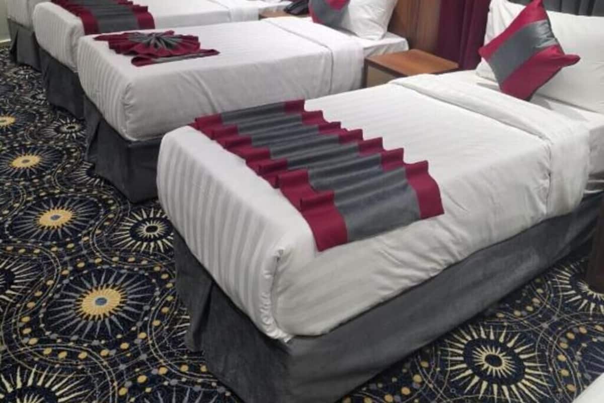 A hotel room with three beds, each with white linens and red accents, a patterned carpet, and an air conditioning unit on the wall.