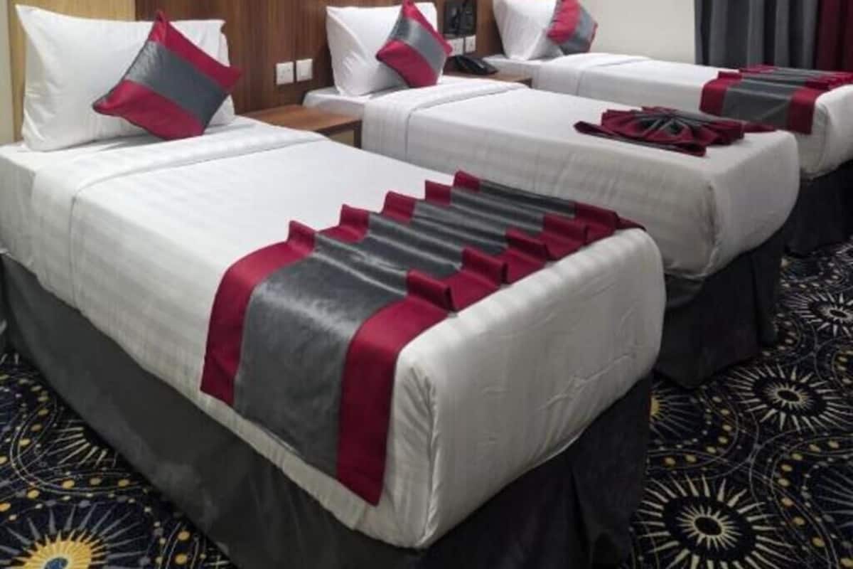 A hotel room with three beds, each with white bedding and red accents. There is a wooden headboard, a painting on the wall, and a patterned carpet.