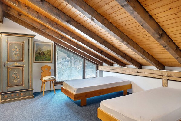 Una habitación con techo inclinado, vigas de madera, una cama, una silla y un armario con motivos florales.
