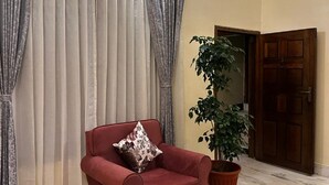 A living room with a red armchair, a wooden coffee table, a large potted plant, and a door in the background. - Heritage Homestay  (Sivasagar)