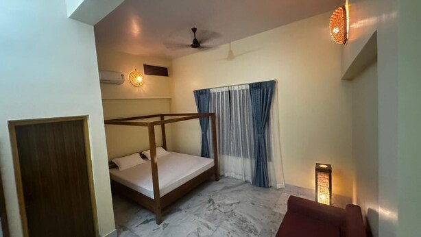 A bedroom with a wooden bed, a ceiling fan, a wall-mounted light, and a window with curtains. - Heritage Homestay  (Sivasagar)