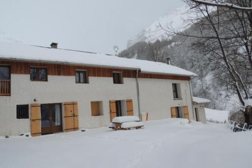 Een gebouw met twee verdiepingen, houten luiken en een sneeuwbedekte nok, omgeven door een besneeerd landschap en bergen.