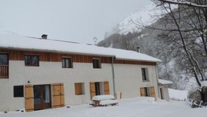 A two-story building with wooden shutters and a snow-covered roof, surrounded by a snowy landscape and mountains. - Gîtes de la Lame - L'Écurie : Authentic charm and serenity in the heart of the Écrins mountains (Réallon)
